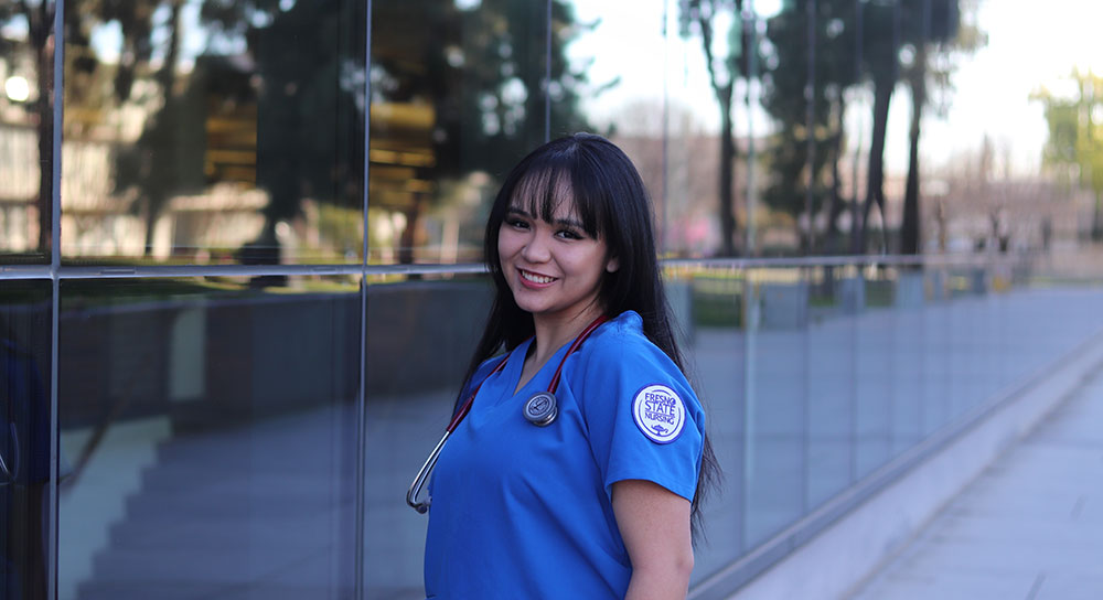 A young Latina woman in a blue shirt smiles in front of a building with reflective windows.