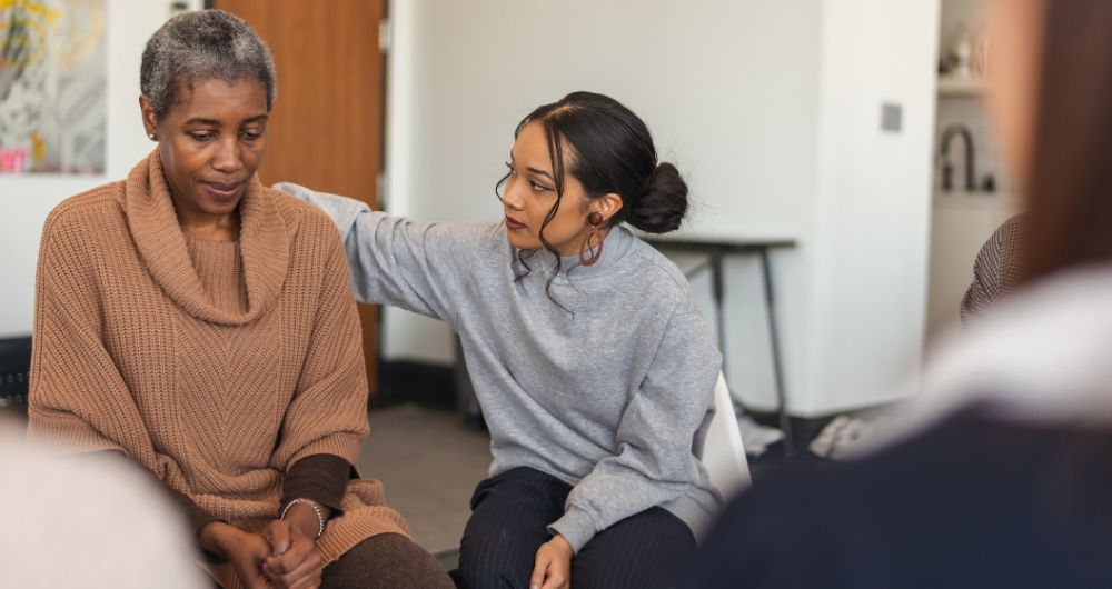 An older Black woman is comforted by a younger Black woman in a support group circle.