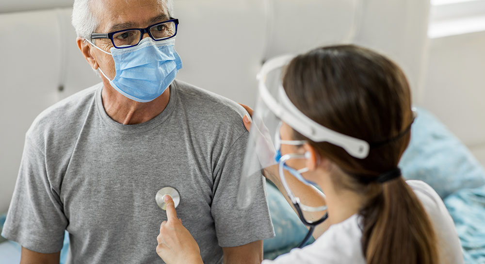 Older man in a mask sits on an exam table as a female doctor places stethoscope on his chest