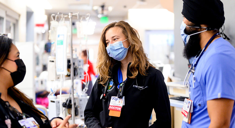 Three masked medical residents talk in a hospital hallway