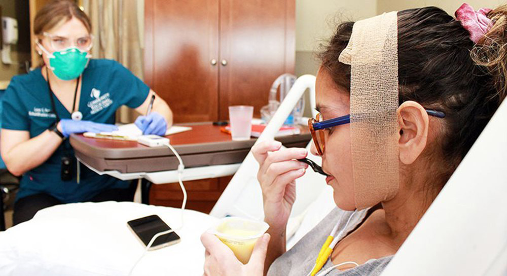 A woman in a hospital bed gingerly eats from a plastic cup