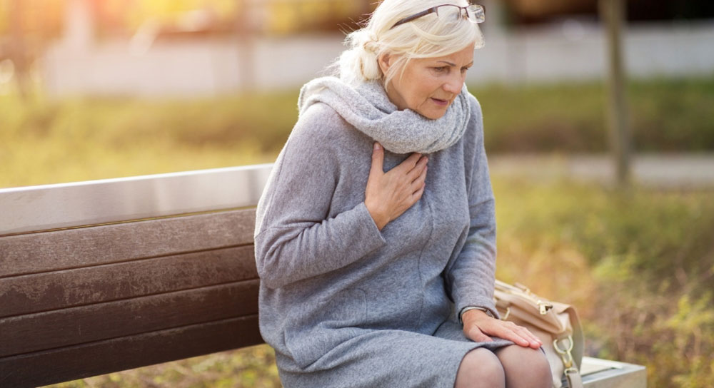 An older white woman sits on a bench and clutches her chest