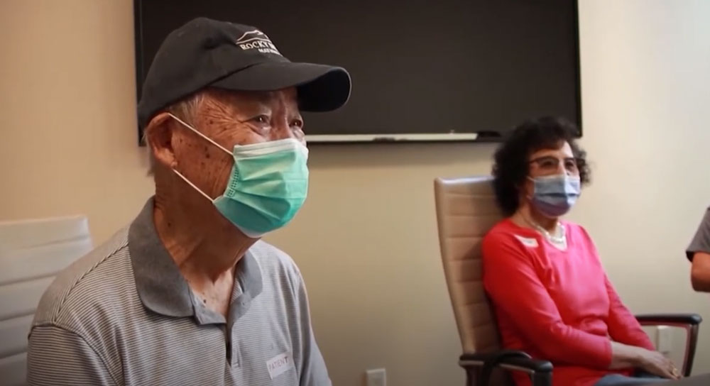 A man in a mask sits next to his wife in a doctor's office