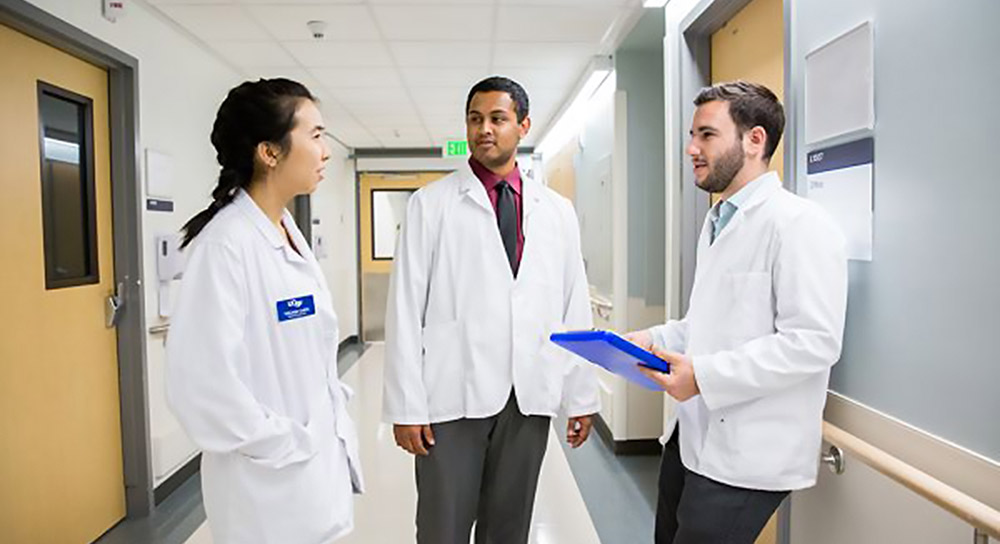 Three students in white coats converse in a hallway