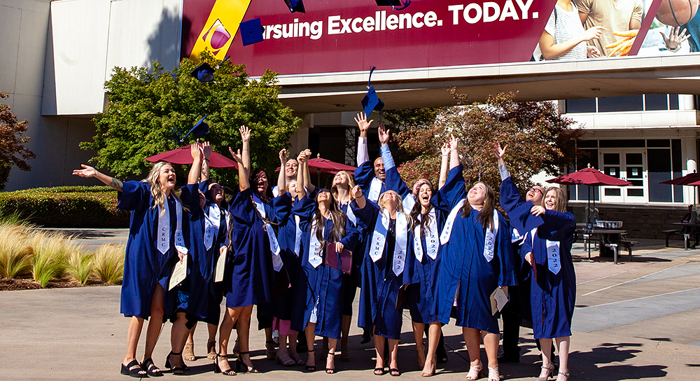 A group of a dozen or so students in graduation gowns toss their caps into the air