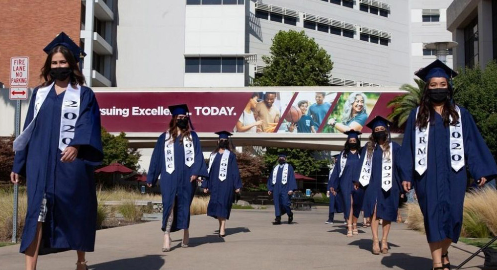 A staggered group of graduates wearing masks march outside
