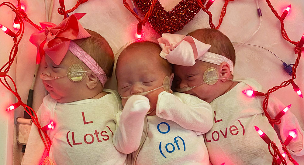 Triplet newborns surrounded by pink lights wear Valentine's clothing