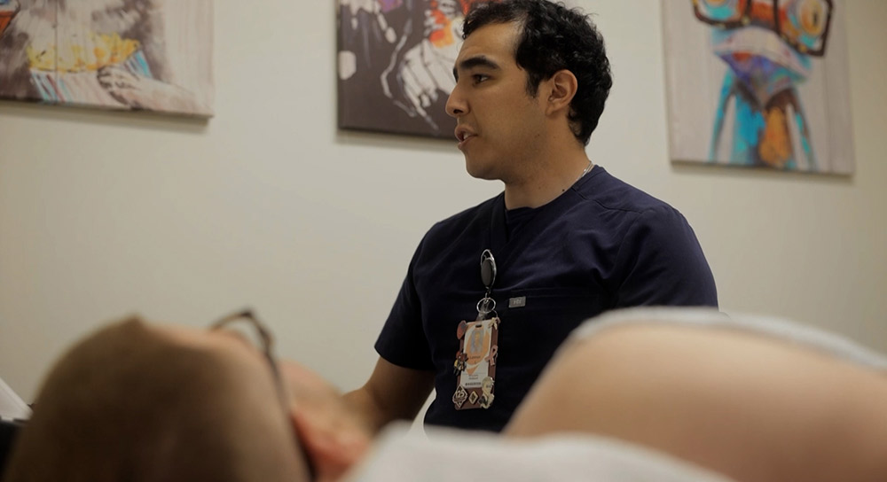 A male sonographer looks at a screen while examining a male patient on an exam table