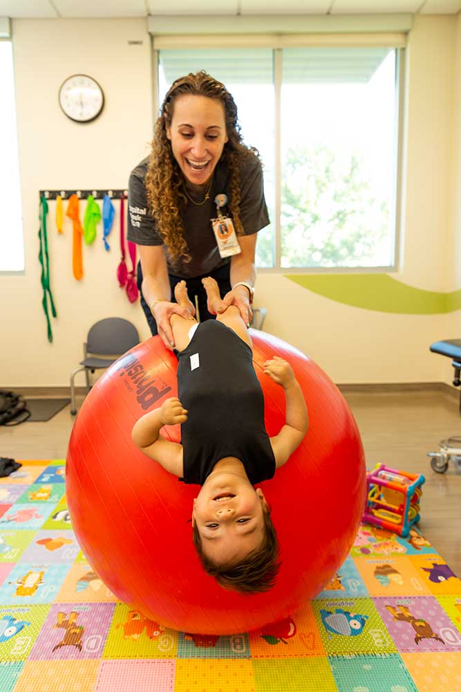 Un trabajador de la salud sostiene a un niño pequeño por las piernas mientras se recuesta sobre una pelota inflable gigante. Se encuentran en una colorida sala de rehabilitación adaptada a los niños.