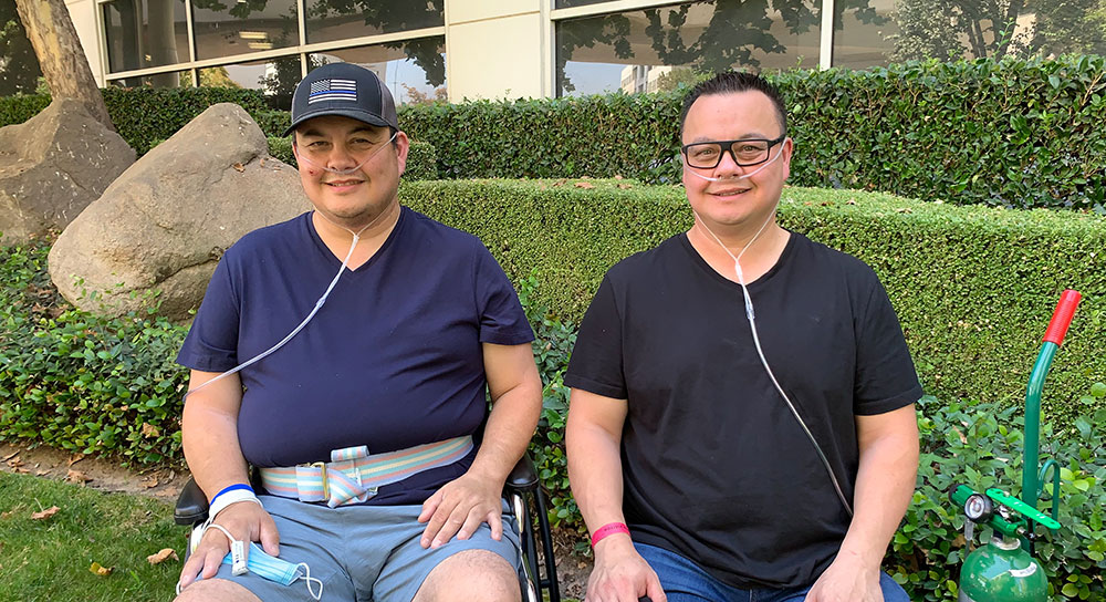 Identical twin men sit side by side, each with oxygen tanks