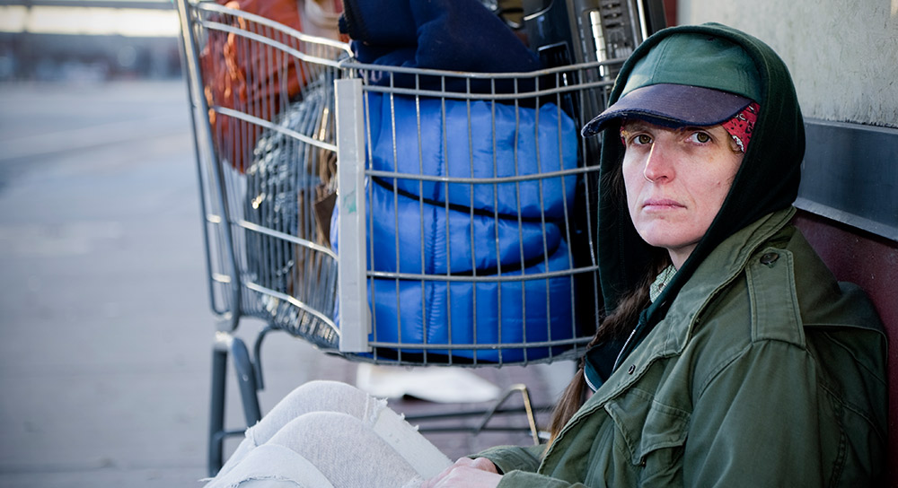 A person sits on the street next to a shopping cart