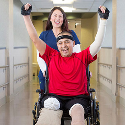 A nurse pushes a man in a wheelchair down a hallway. He has his arms raised in victory