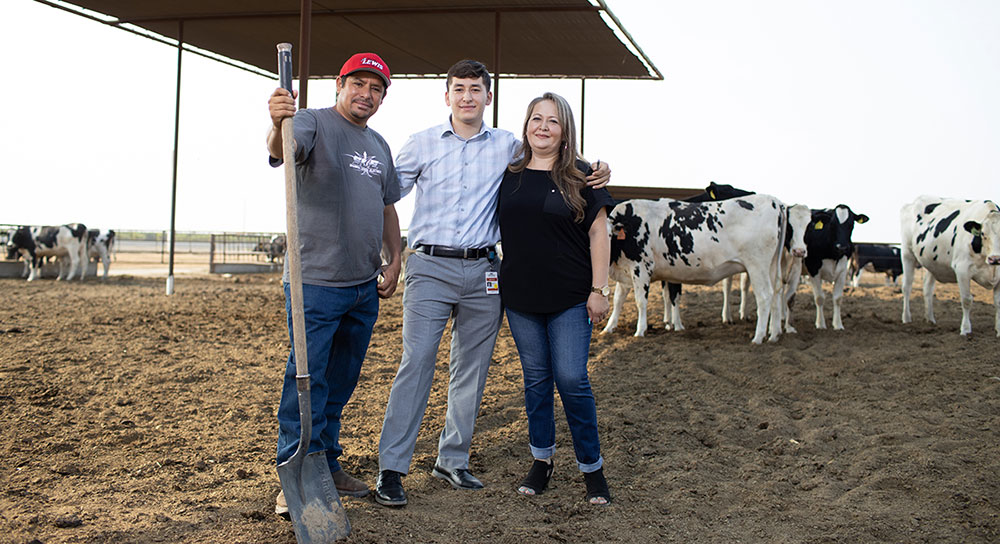 A college-aged man stands in between his mother and father with cows in the background