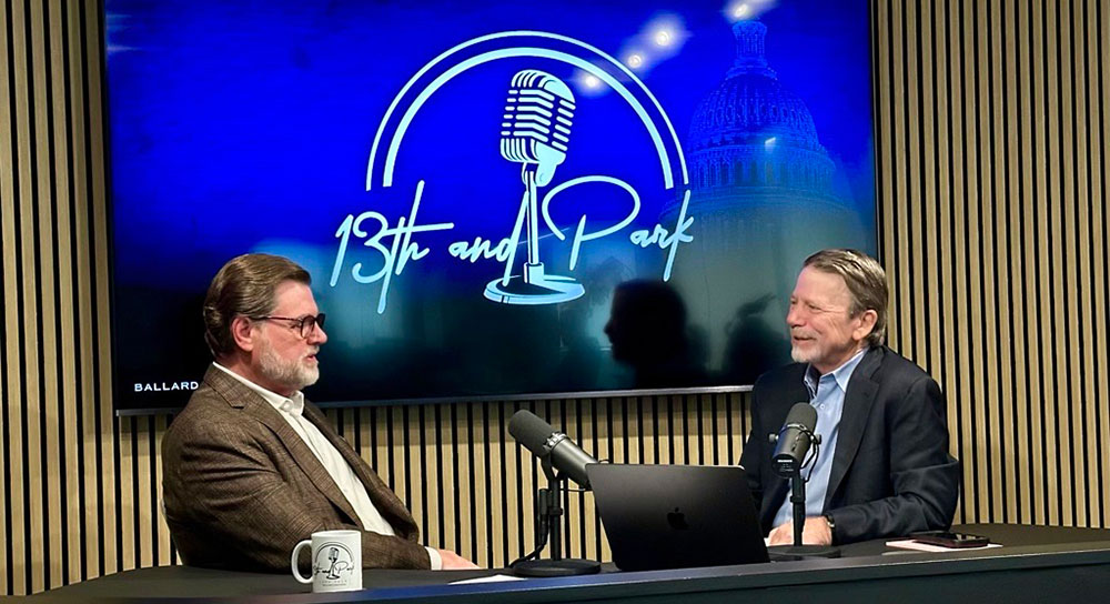Two men in suits sit in front of microphones. A screen with the logo reading 13th and Park is behind them.