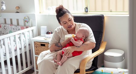 a Latina woman feeds her crying baby from a bottle as she sits in a chair in her baby's room