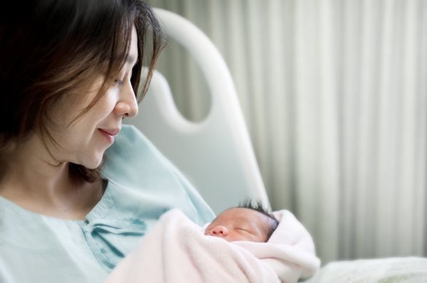 Joven madre de origen asiático que mira con amor a su bebé recién nacido que tiene en brazos mientras está recostada en la cama de un hospital.