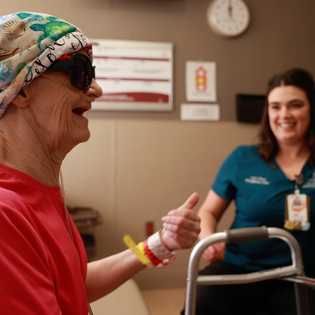 A female patient in a head wrap smiles as her rehab therapist smiles in the background