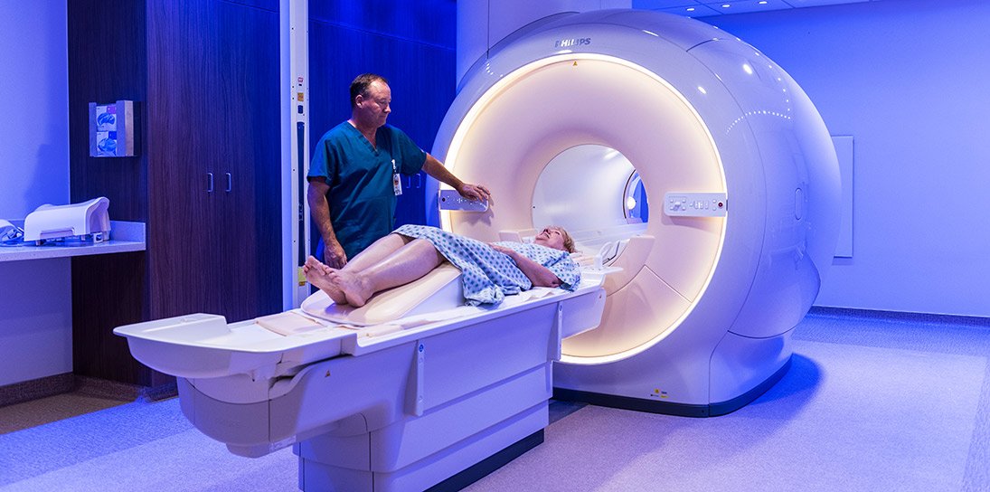 Paciente femenina de unos 65 años preparándose para que la examinen en una máquina grande de escaneo. Técnico médico de pelo marrón con uniforme hospitalario verde azulado parado ante la paciente.