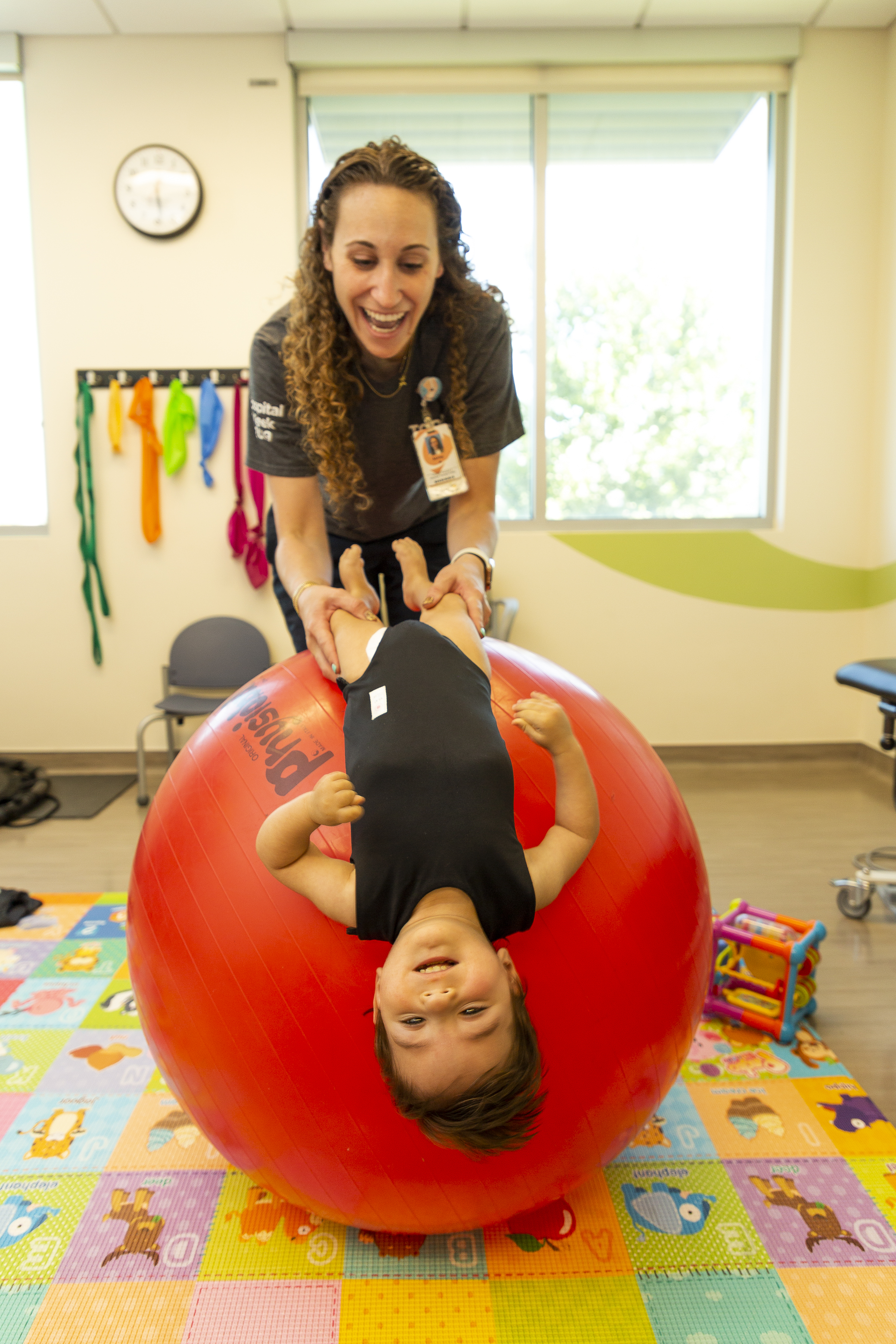 Un trabajador de la salud sostiene a un niño pequeño por las piernas mientras se recuesta sobre una pelota inflable gigante. Se encuentran en una colorida sala de rehabilitación adaptada a los niños.