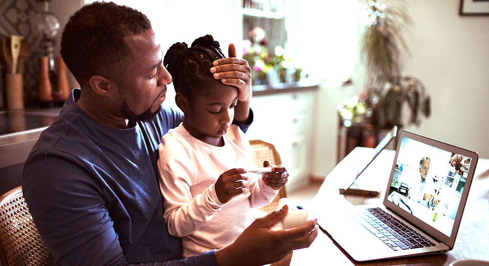 A father (Black) and his young daughter (also Black) sit on front of their laptop computer while a doctor (male, white) speaks on health matters. 