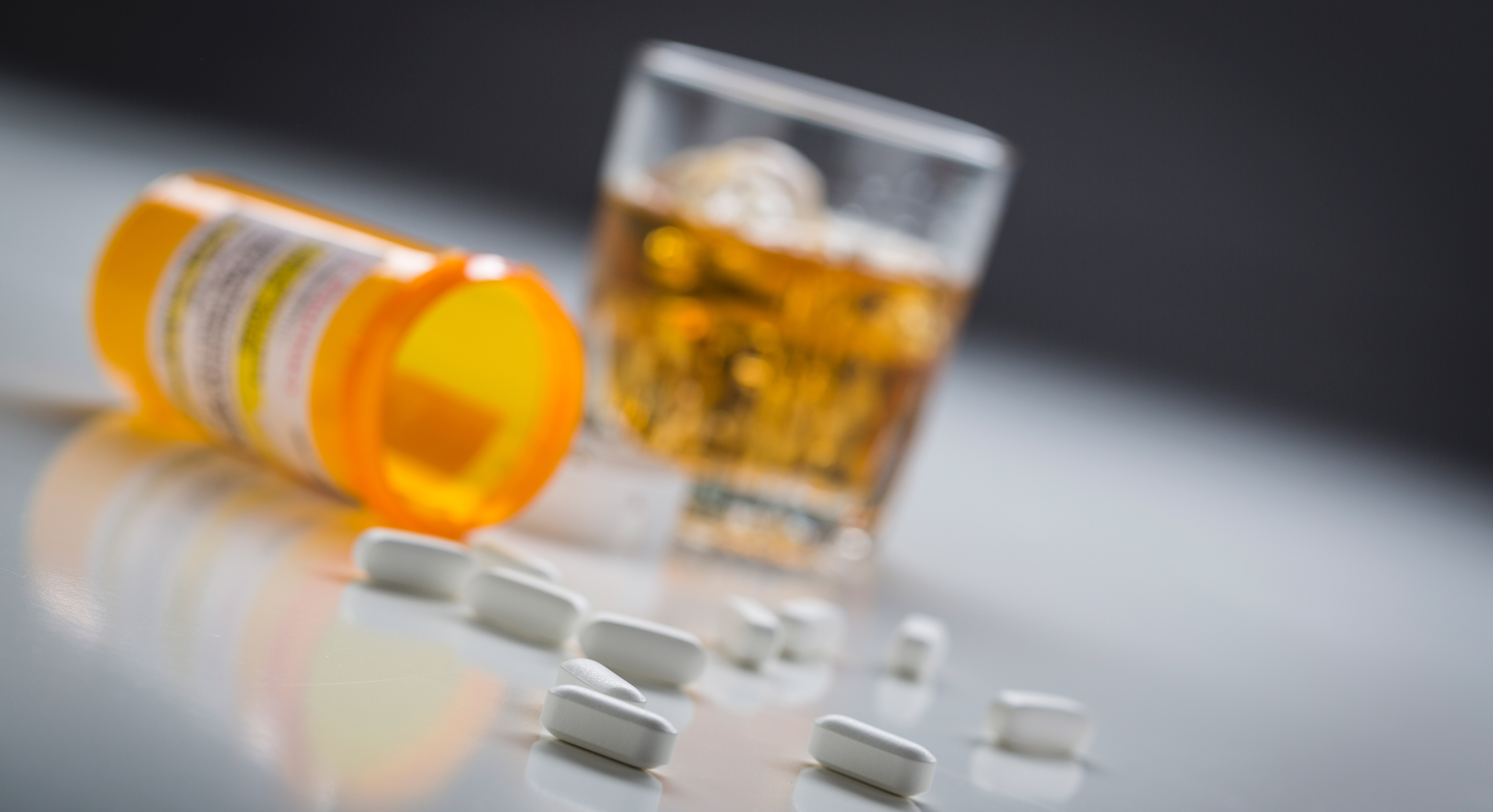 A pill bottle with scattered pills rests on a table next to a glass of whiskey