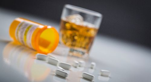 A pill bottle with scattered pills rests on a table next to a glass of whiskey