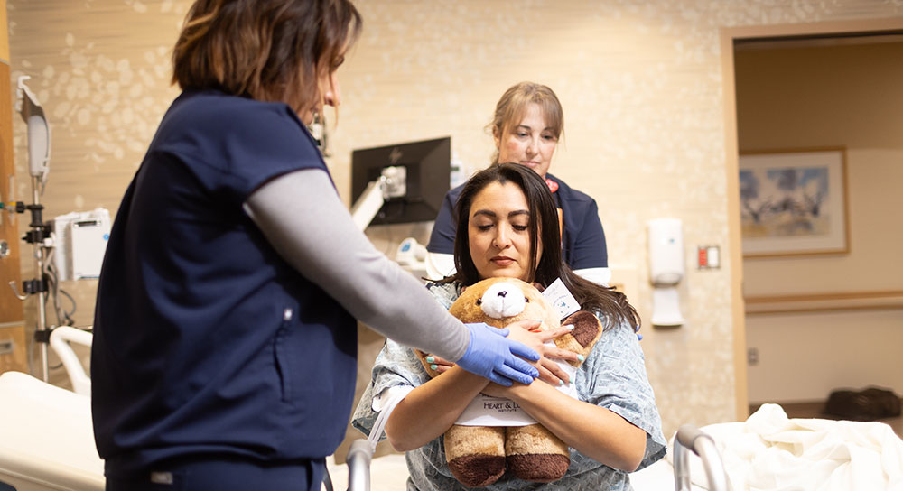 A physical therapist in scrubs holds up a stuffed bear for a patient in a wheelchair to grasp.