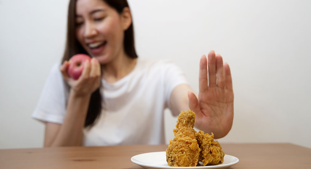 A young Asian woman, pretends to take a bite of an apple while her handing out to block a piece of fried chicken.