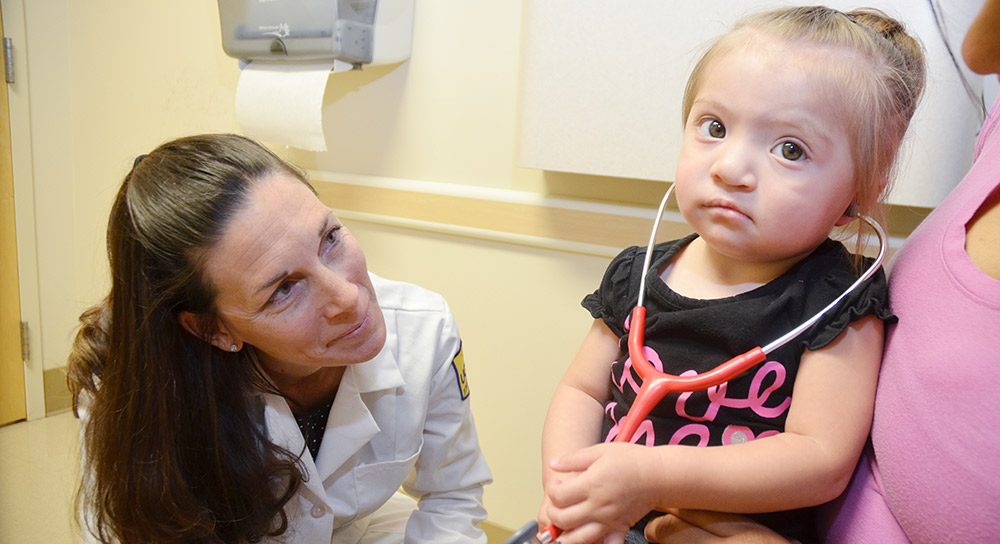 Una médica blanca de UCSF se inclina para sonreír a una niña pequeña de origen hispano