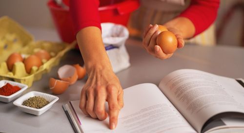 View of a woman's hands as she skims a recipe book with one finger while holding a brown egg in the other hand. 