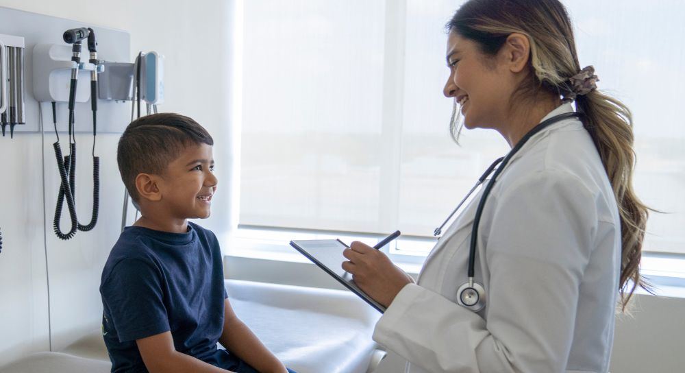 A pediatric patient, male, smiles at his doctor as she writes a note in his chart