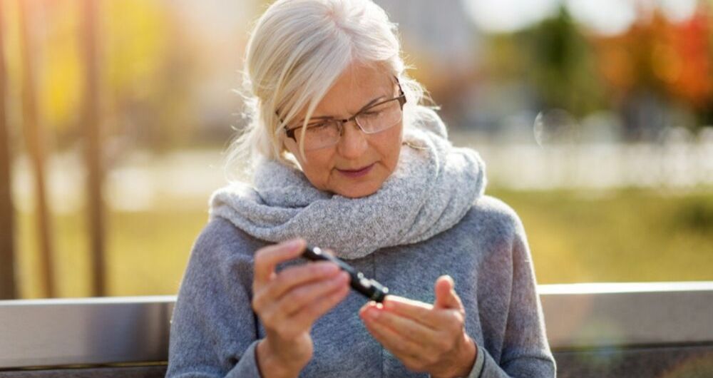 An older white woman with white hair sits outside on a bench and pricks her finger with 