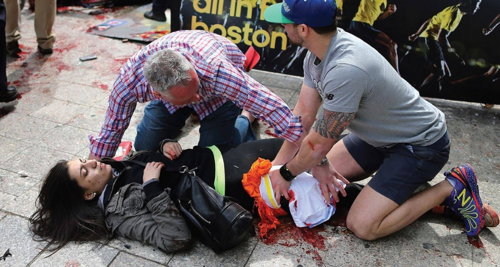 A young woman lays ont he ground bleeding while two men attend to her injuries