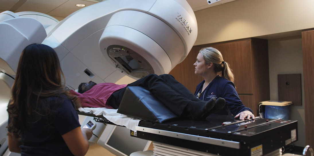 A young Indian man in a maroon shirt reclines as two female medical technicians, one with brown hair and one with blond hair, prepare him for assessment by a white imaging machine.