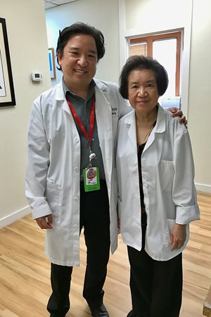 Dr. New Sang (male) stands next to his mother, Dr. Orathai Sangrujiveth. Both are wearing white doctor's coats and are standing in a hospital hallway.