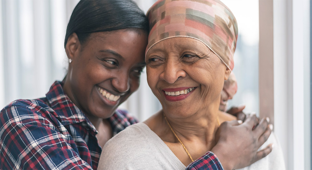 A young woman with dark skin hugs an older woman with lighter skin who is wearing a headscarf. They are both smiling.