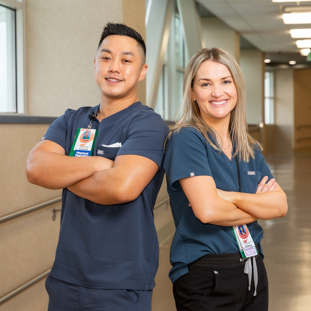 A young male healthcare worker and a young female healthcare worker stand back to back in a hospital hallway.