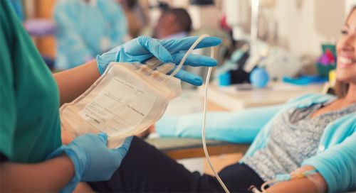 Close up of an empty blood bag held by gloved hands. In the background, a smiling young Asian woman sits in a chair ready to donate blood.