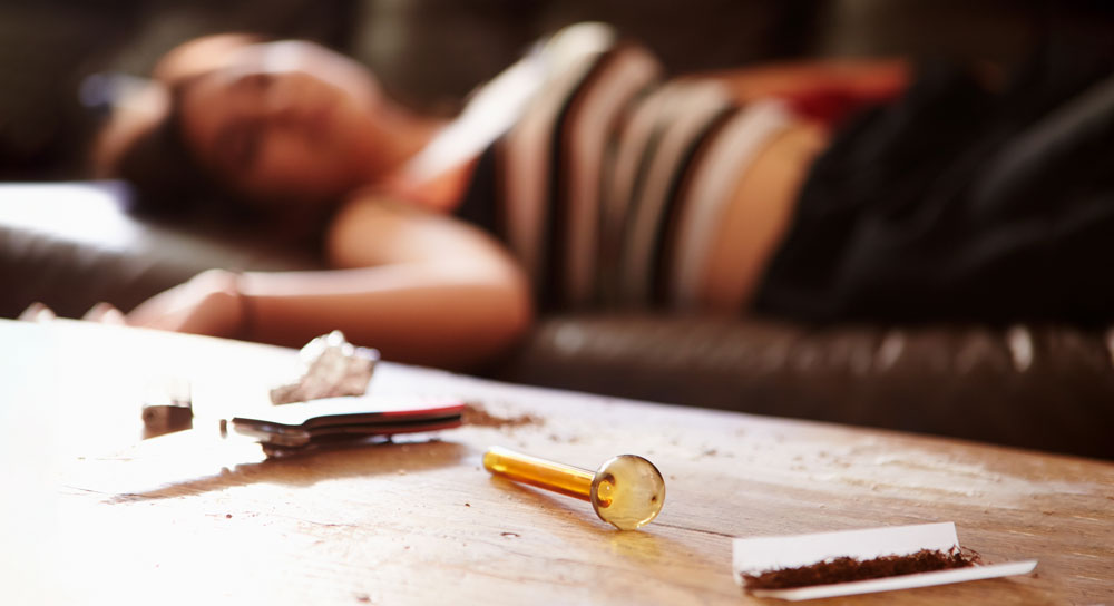 Drug paraphernalia in the foreground as a young woman is lying, blurry, in the background