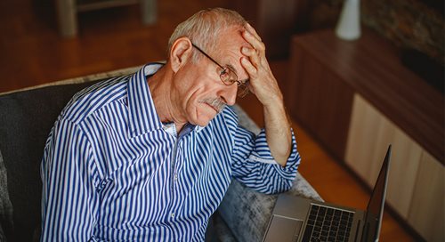 A senior citizen, white and male, rests his head in his hands and looks forlorn. He is wearing a blue and white striped shirt and looking at a laptop computer.