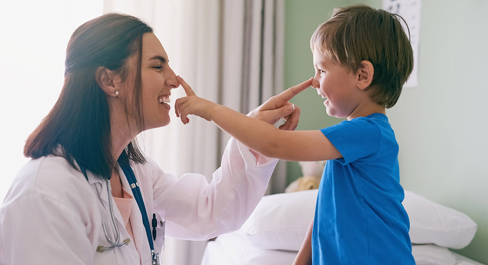 A young female doctor with shoulder-length brown hair playfully boops the nose of a young boy who boops her nose back. 