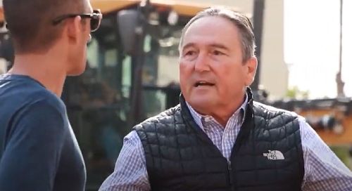 A white man, middle-aged, in a black puffer vest is in conversation with another man whose face is obscured.