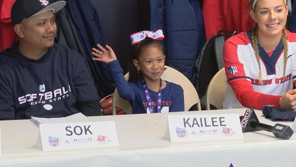 Kailee Sidamrong-Phan waves from a table, sitting next to her father and Fresno State softball players