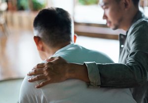 seen from behind: a seated man puts his arm around another in a comforting way