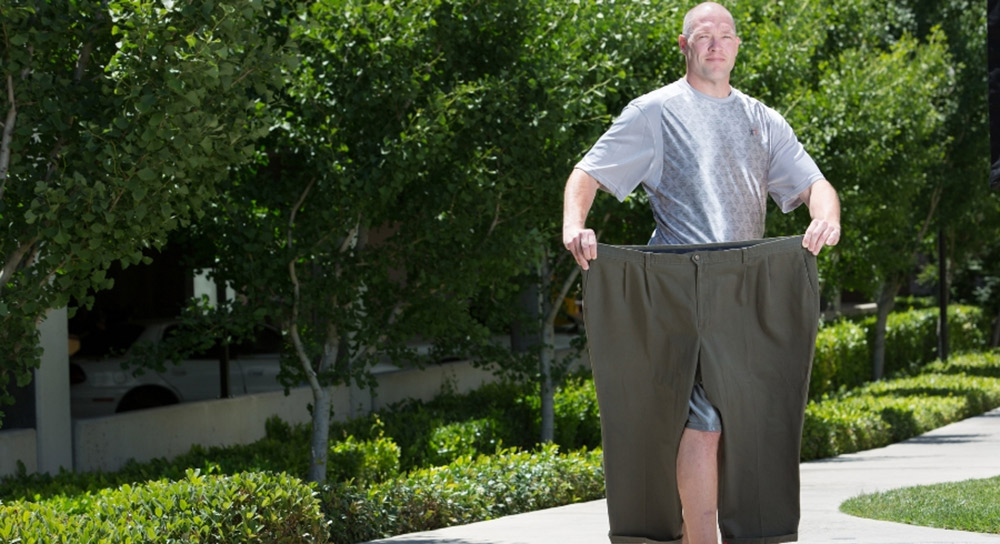 A white man with no hair holds up a very large pair of pants in front of him to illustrate considerable weight loss