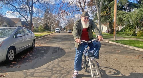 Paul Wilson rides his bike down the street on a sunny day with blue sky and clouds in the background