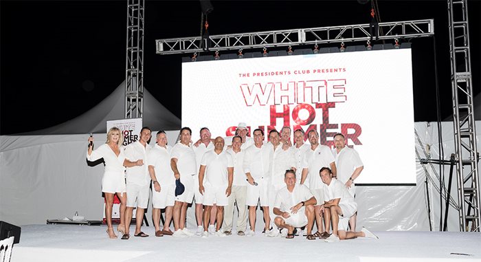 A group of about a dozen men, dressed in all in white, stand on a stage. Behind them a screen reads, "The Presidents Club presents White Hot Summer Night."