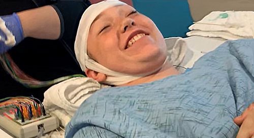 Karmen Reeves sonríe recostada en su cama con una bata quirúrgica y vendas en la cabeza.