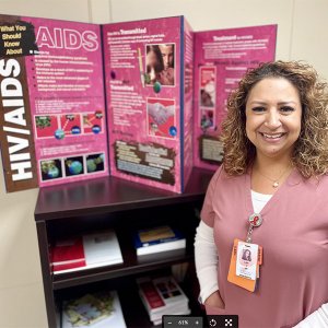 Lupe Vargas, in salmon scrubs, stands next to a display of info about HIV/AIDS