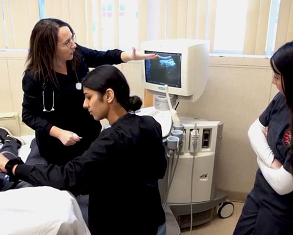 a healthcare worker in scrubs points to a monitor as students look on